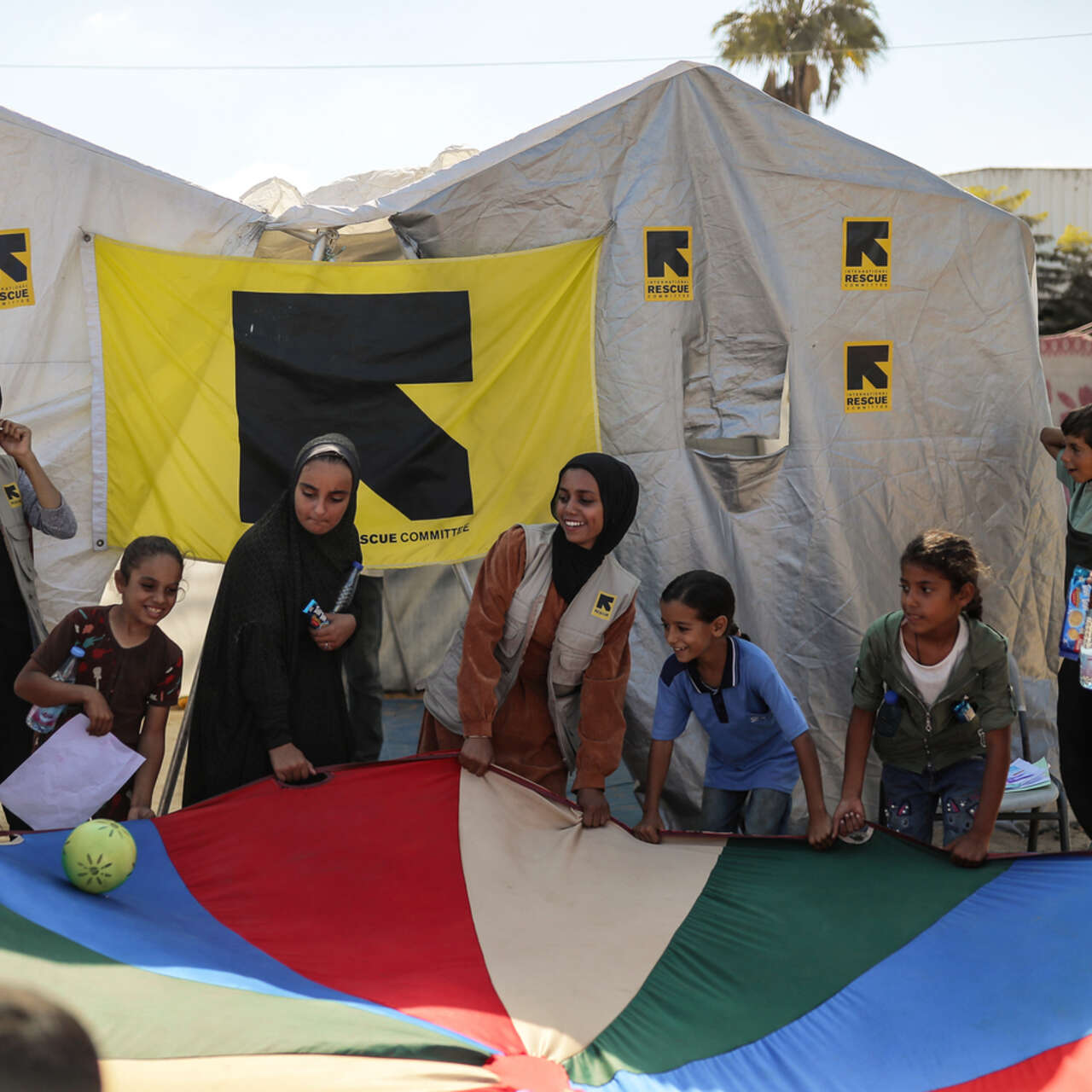 Barn i Gaza och RESCUE:s personal håller tillsammans upp ett stort tygstycke i många färger. I bakgrunden syns tält med RESCUE:s logga.